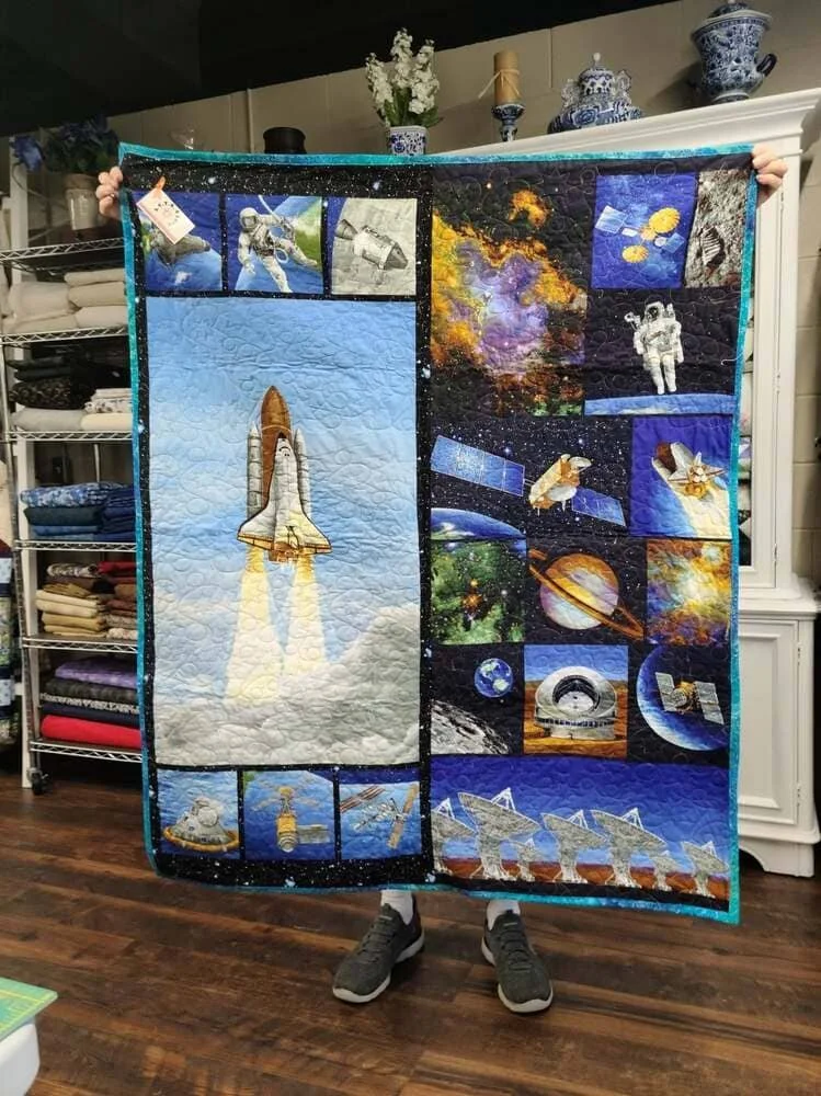 Person holding a space-themed quilt featuring rockets, astronauts, planets, stars, and galaxies in a room with shelves and decorative items.