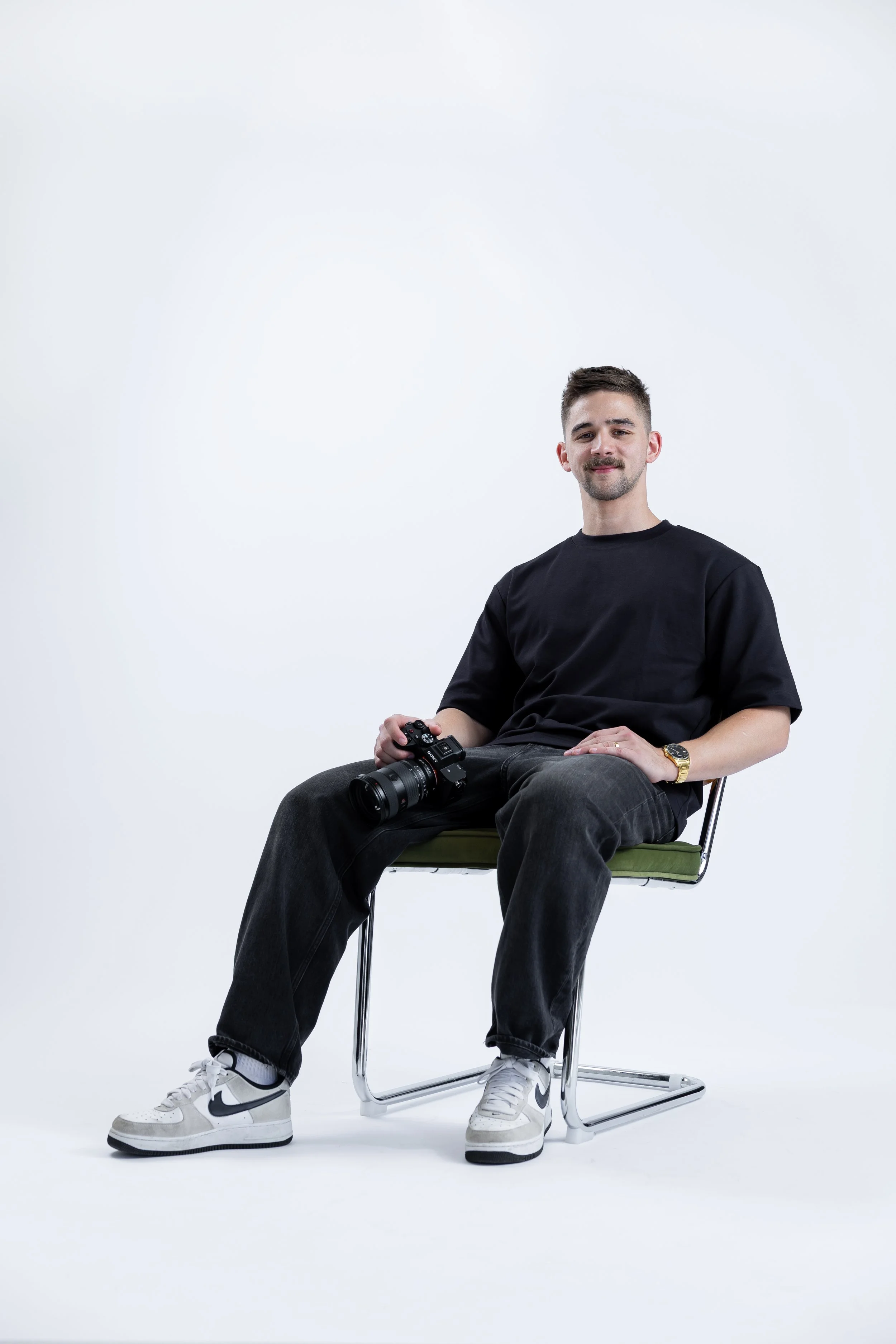 A young man with short brown hair and a goatee, wearing a black T-shirt, black jeans, and white Nike sneakers, sits on a metal and green cushioned chair against a plain white background, holding a camera in his right hand and smiling slightly.