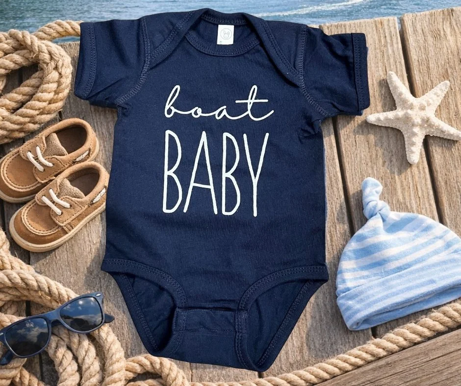 A navy blue baby onesie with the words "boat BABY" written on it, surrounded by a pair of tan baby shoes, a starfish, a striped baby hat, ropes, and a pair of sunglasses on a wooden surface.