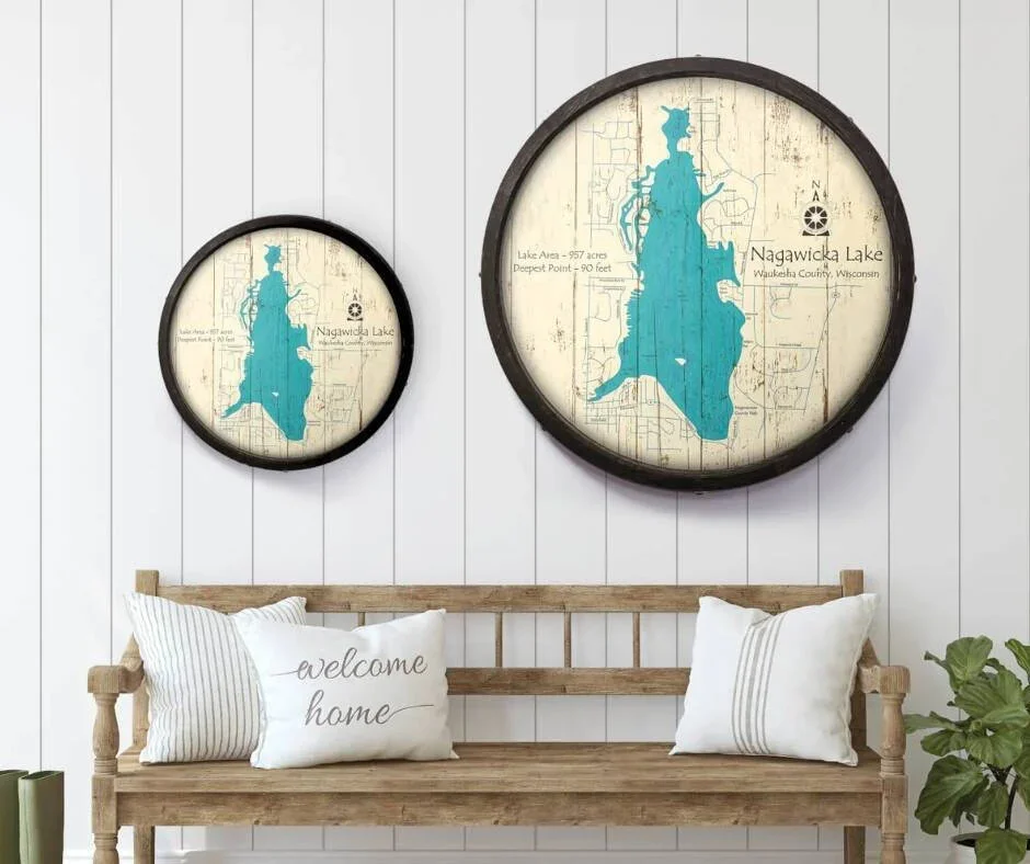 Two round wall signs featuring maps and information about Nagawicka Lake in Wisconsin, hung above a wooden bench with pillows, on a white paneled wall.