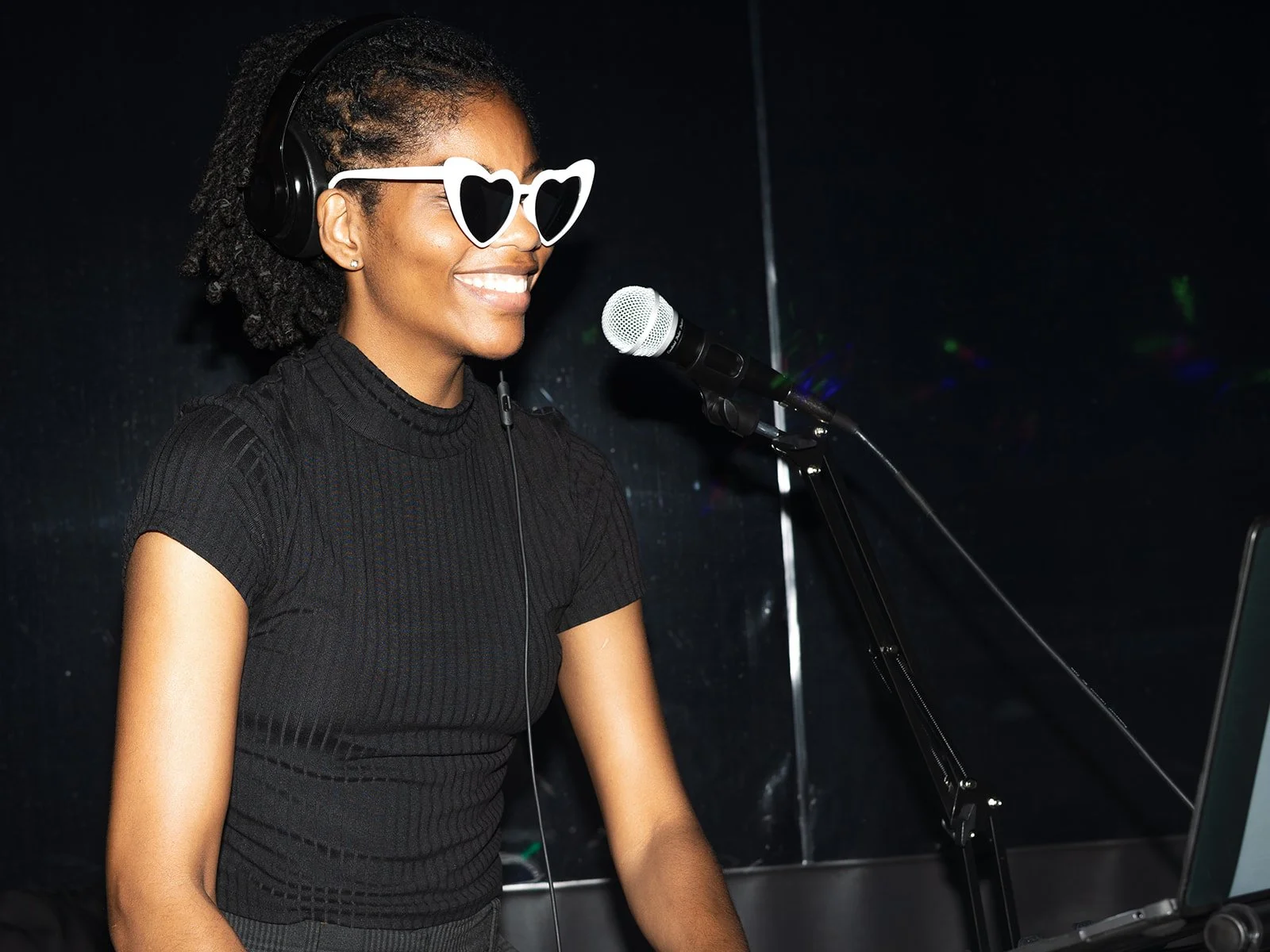 A woman wearing heart-shaped sunglasses, headphones, and a black top, smiling while singing or speaking into a microphone in a dark environment.