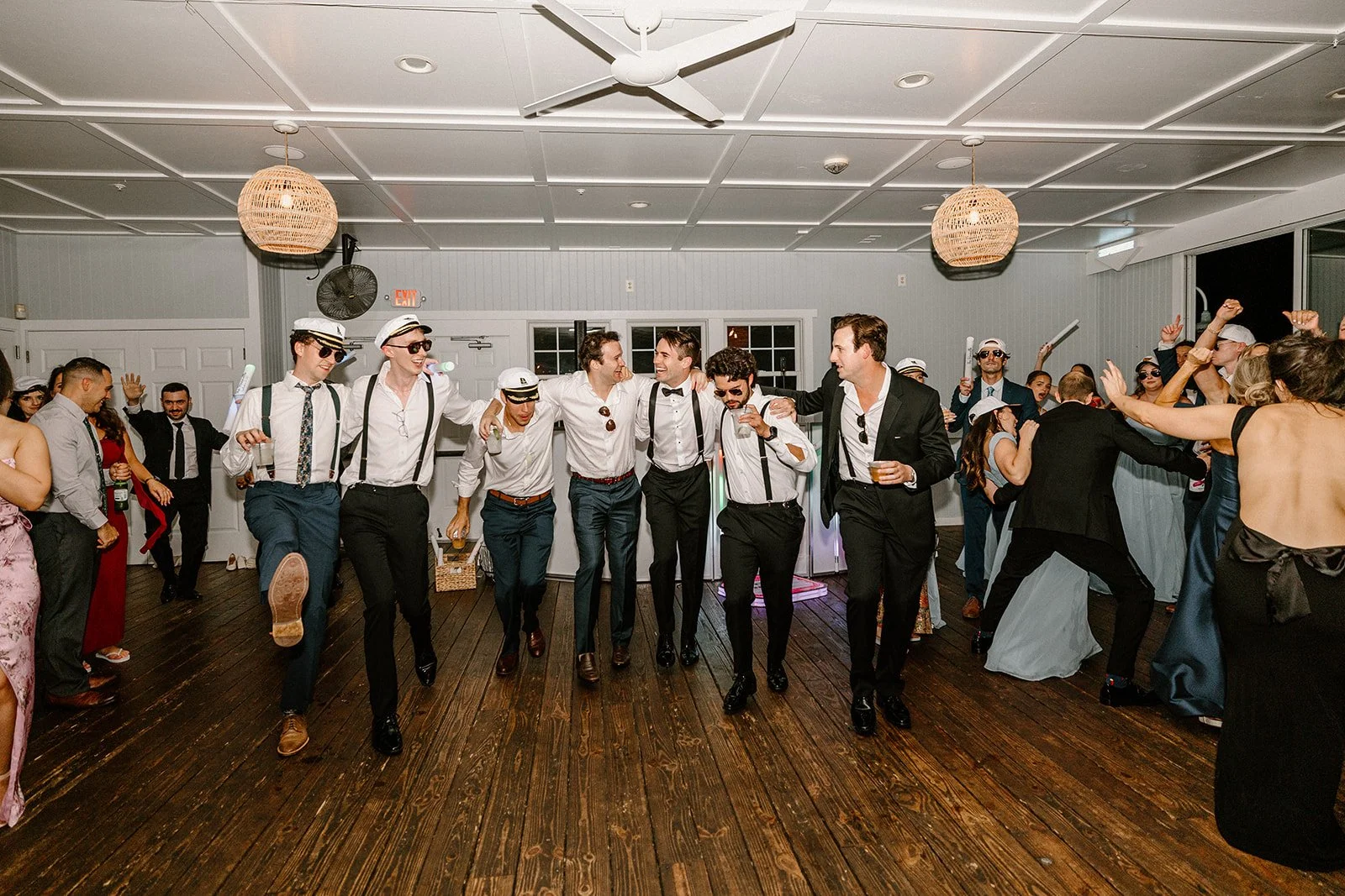 Group of people dancing and celebrating at a party, some dressed in vintage style with white shirts, suspenders, and sailor hats, others in formal attire with tuxedos, in a decorated indoor venue with wooden floors and ceiling fan.