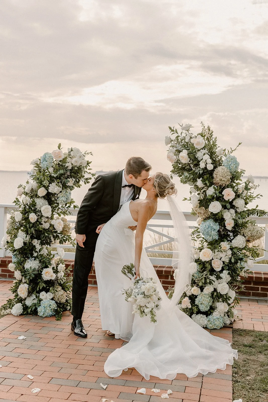 A newlywed couple sharing a kiss during their wedding ceremony at an outdoor location with floral arrangements on either side and a scenic view in the background.