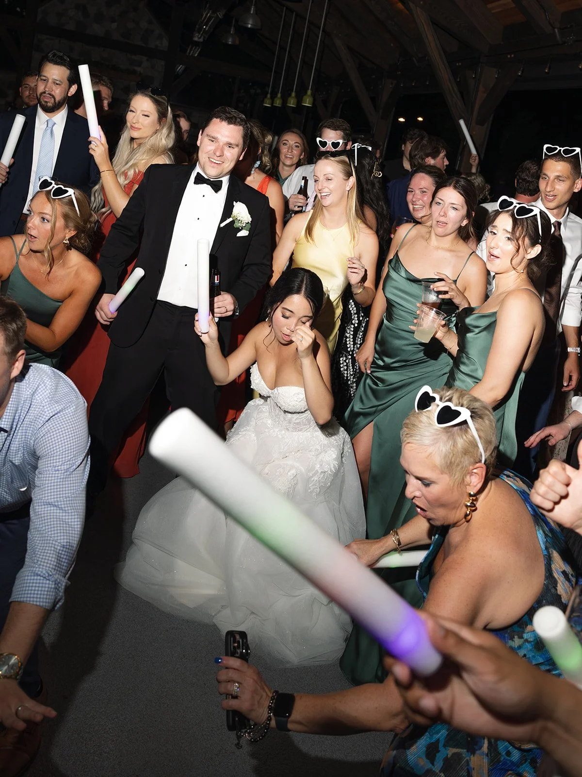 Group of people at a wedding reception, some holding glow sticks, wearing party accessories like heart-shaped sunglasses, and dressed in formal attire, including the bride in a white gown and the groom in a tuxedo.