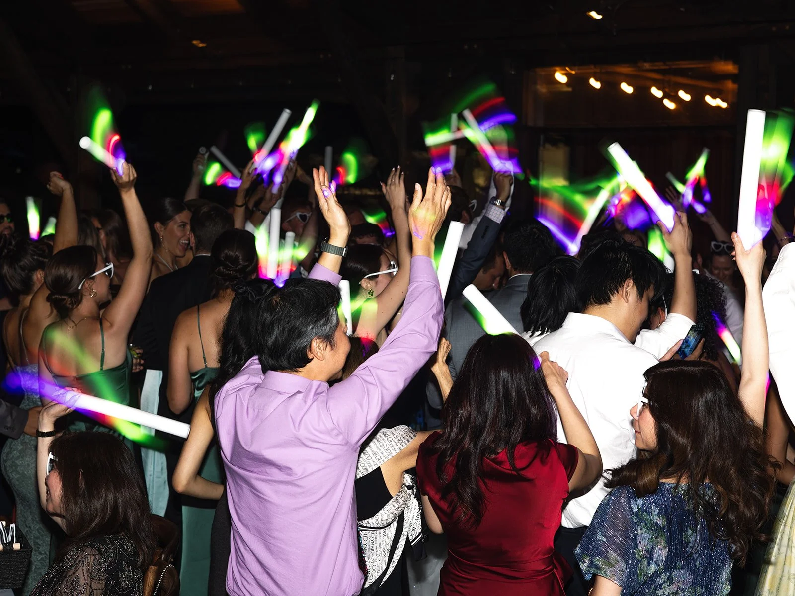 Crowd of people dancing at a party, holding glow sticks and wearing evening attire, with colorful lights in the background.