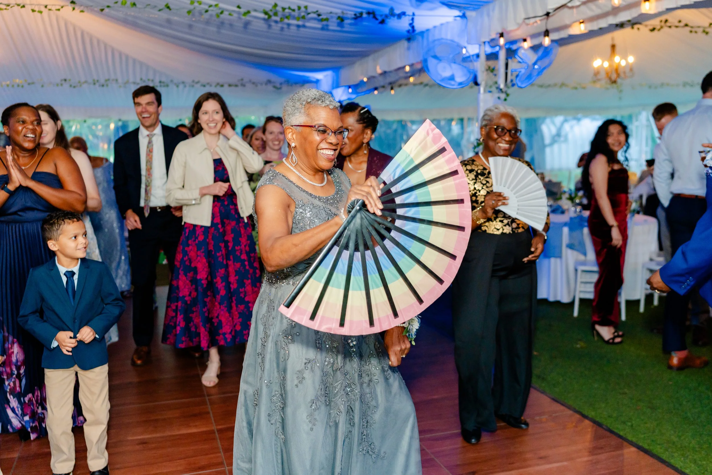 A group of people celebrating at an event, with women holding paper fans and smiling under a decorated tent with string lights.