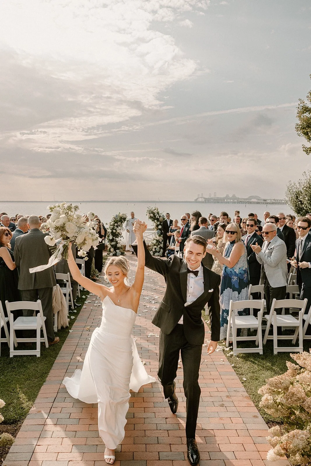 A bride and groom walk down the aisle outdoors by a body of water, smiling and holding hands, with guests clapping and celebrating in the background.