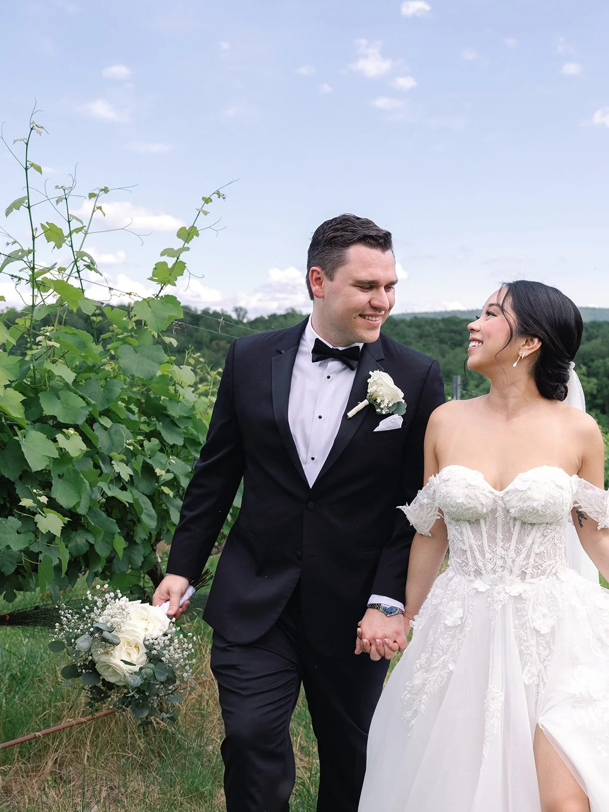 A groom and bride holding hands and walking through a vineyard, smiling at each other. The groom is wearing a black tuxedo with a white shirt and black bow tie, holding a bouquet of white roses. The bride is in a white off-shoulder wedding dress with