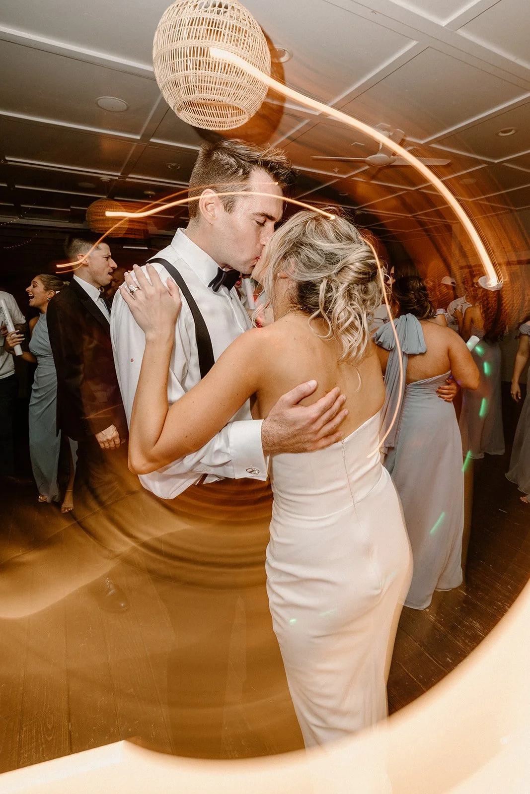 A couple sharing a dance close together at a wedding, surrounded by other guests on the dance floor. The man is wearing a white shirt with suspenders and a bow tie, and the woman is in a strapless white dress with blonde, curly hair. They are dancing
