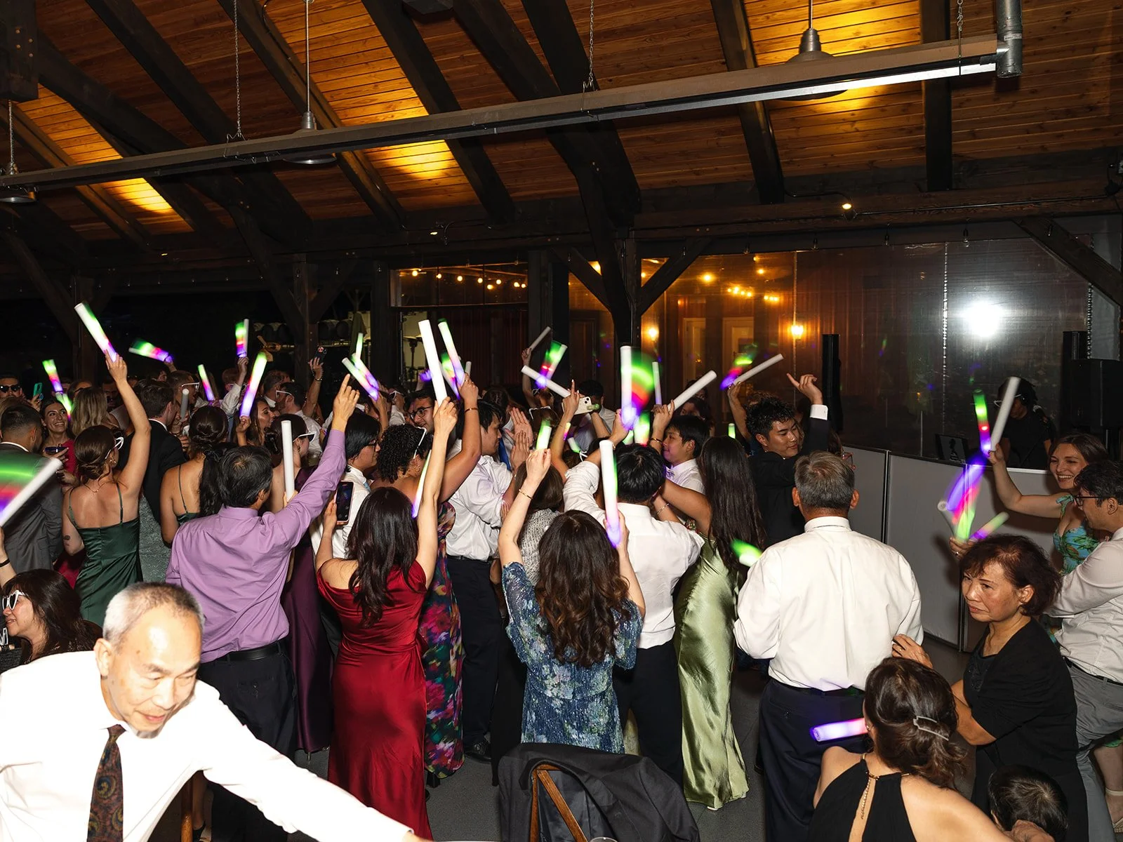 Crowd of people at a party, dancing and celebrating with glow sticks at an indoor venue with wooden ceiling.