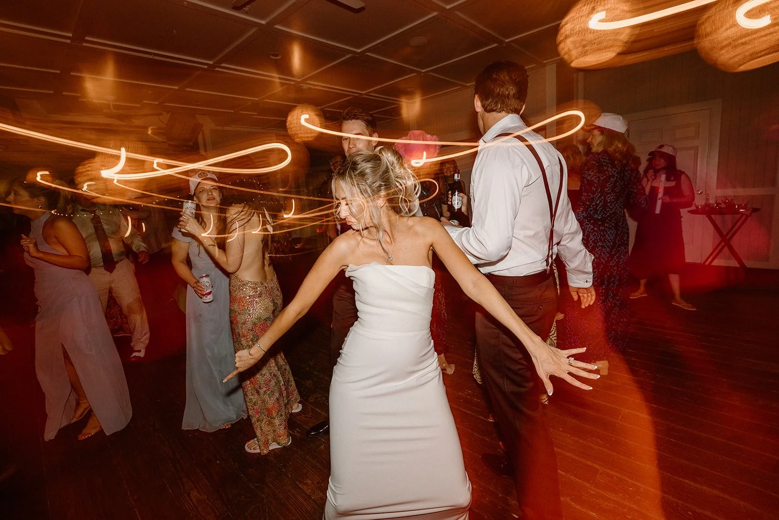 People dancing at a wedding reception, with a bride in a white strapless dress in the center and others in the background holding drinks.