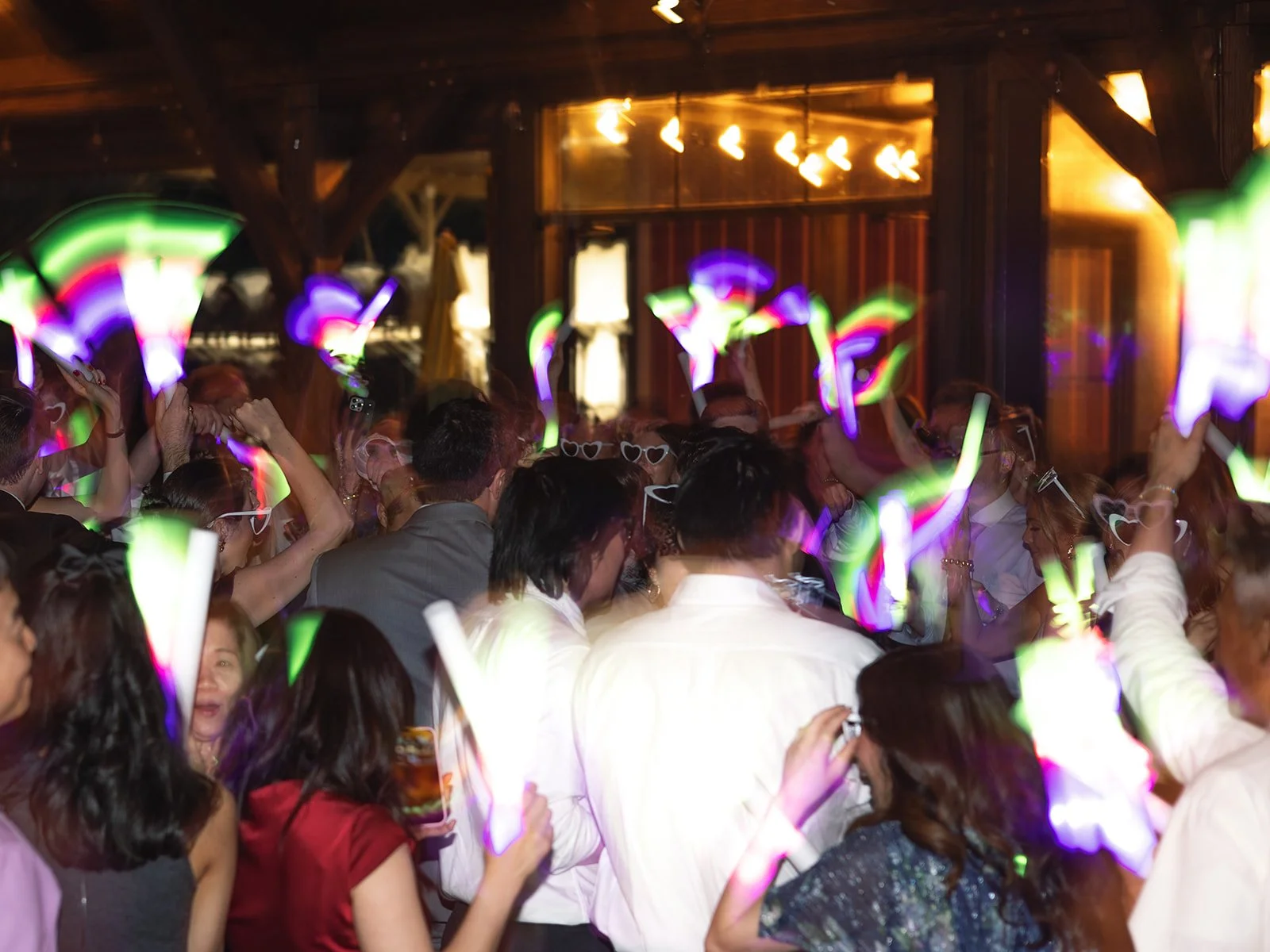 A crowded dance floor at a party with colorful glow sticks and people dancing under warm overhead lighting in a rustic venue.