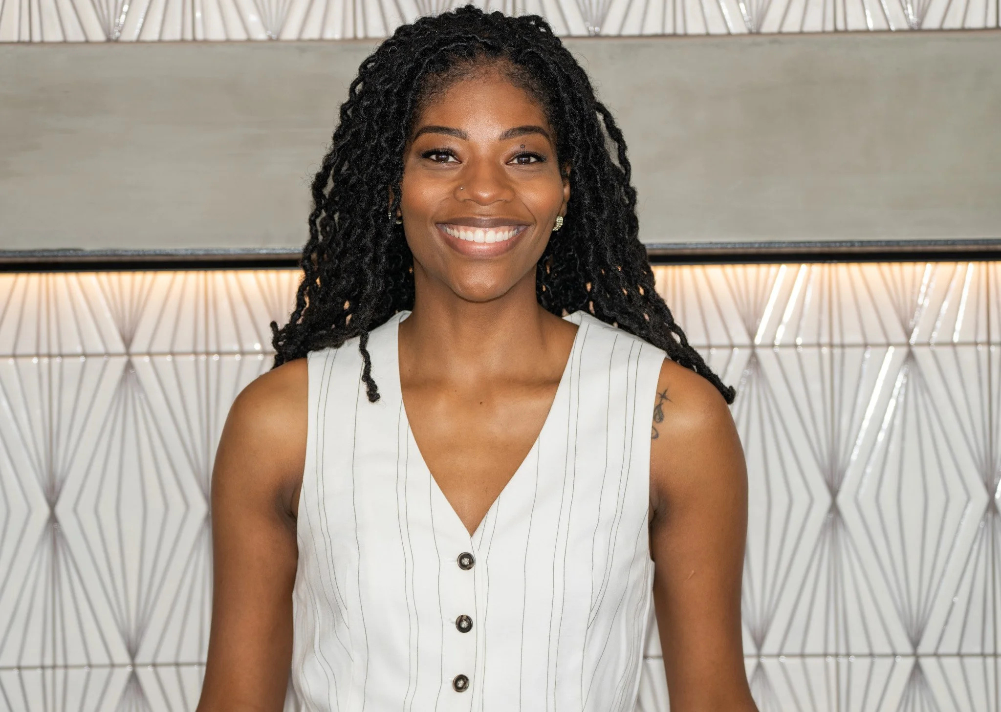 A woman with long locs hair  smiling, wearing a sleeveless white top with vertical stripes, standing in front of a geometric patterned wall.