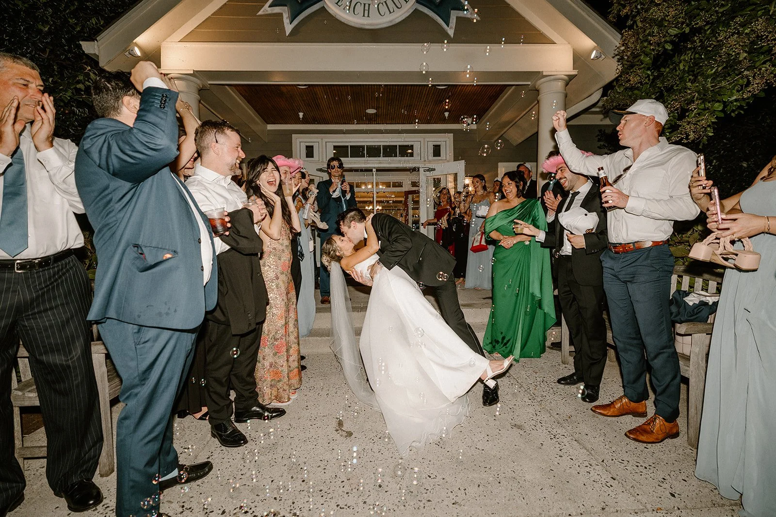 A newlywed couple kissing and dancing at their wedding reception, surrounded by guests celebrating and taking photos outside a building.