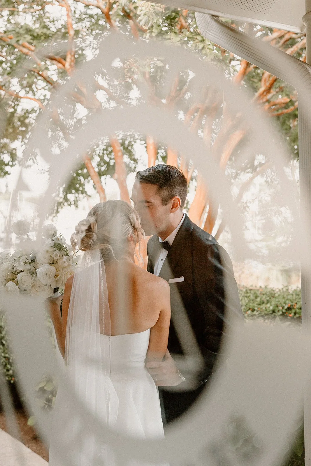 A bride and groom sharing a kiss during their wedding ceremony outdoors, with trees in the background, viewed through a decorative mirror frame.