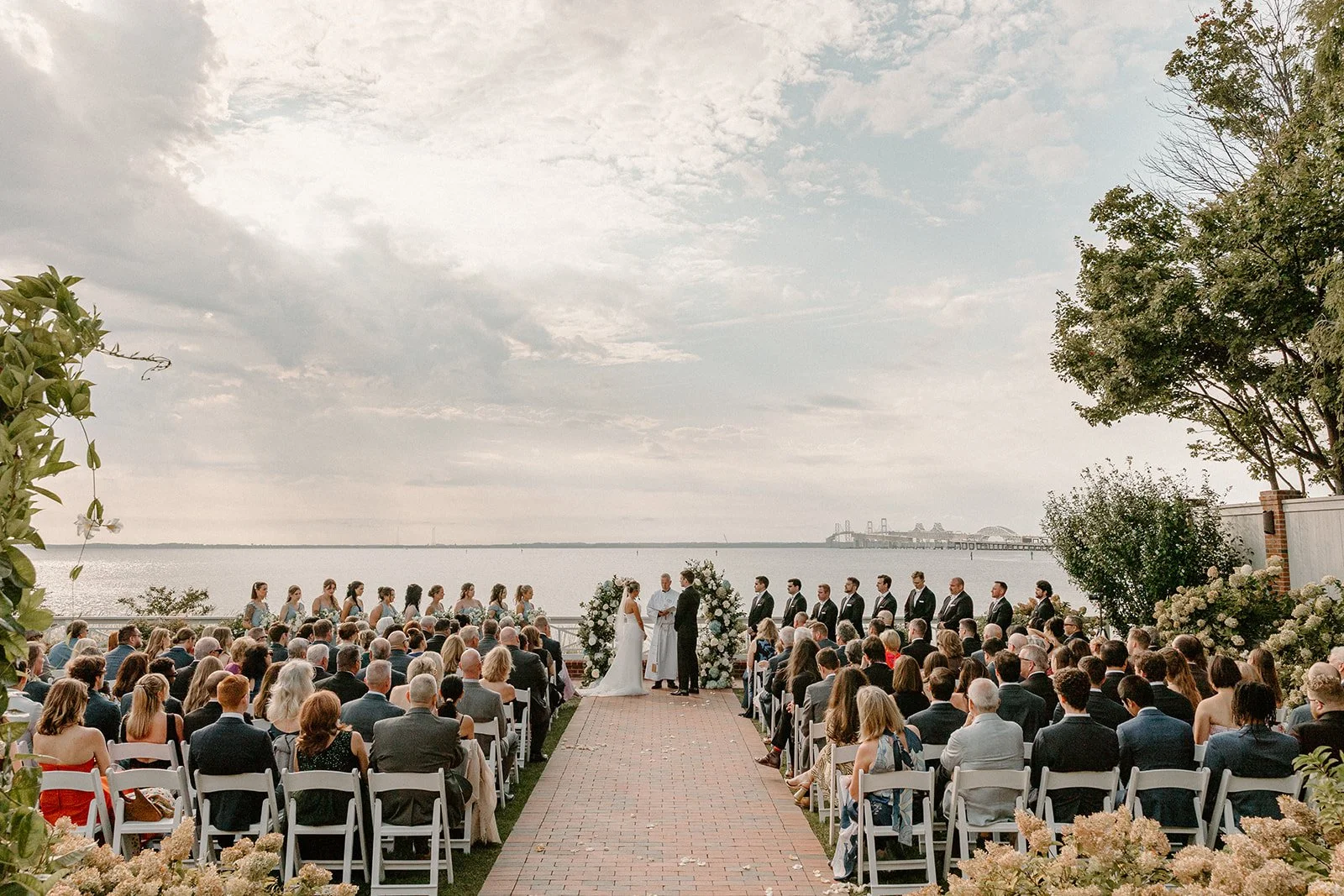 Outdoor wedding ceremony by a waterfront with a large gathering of guests, a couple exchanging vows, and a floral arch. Overcast sky with some clouds, trees and distant structures visible across the water.