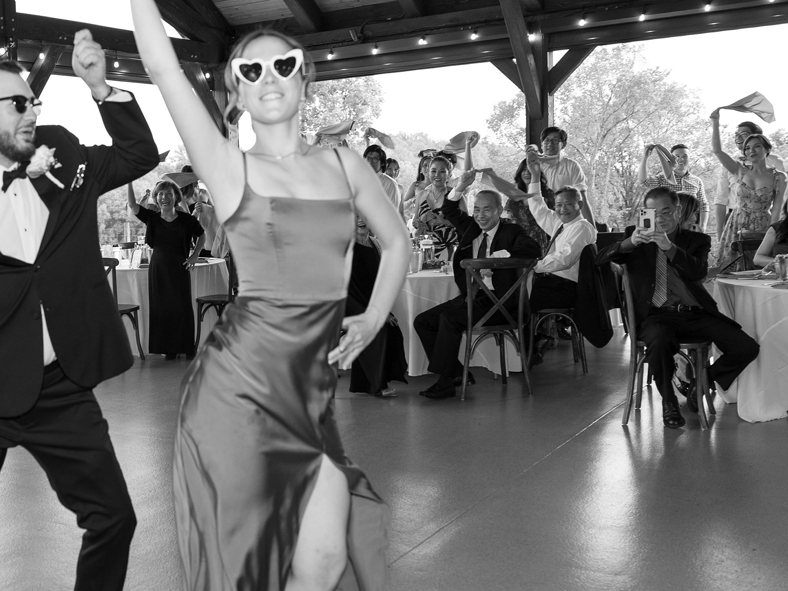 People dancing at a wedding celebration in a decorated venue with tables and guests watching, some taking photos, in black and white.