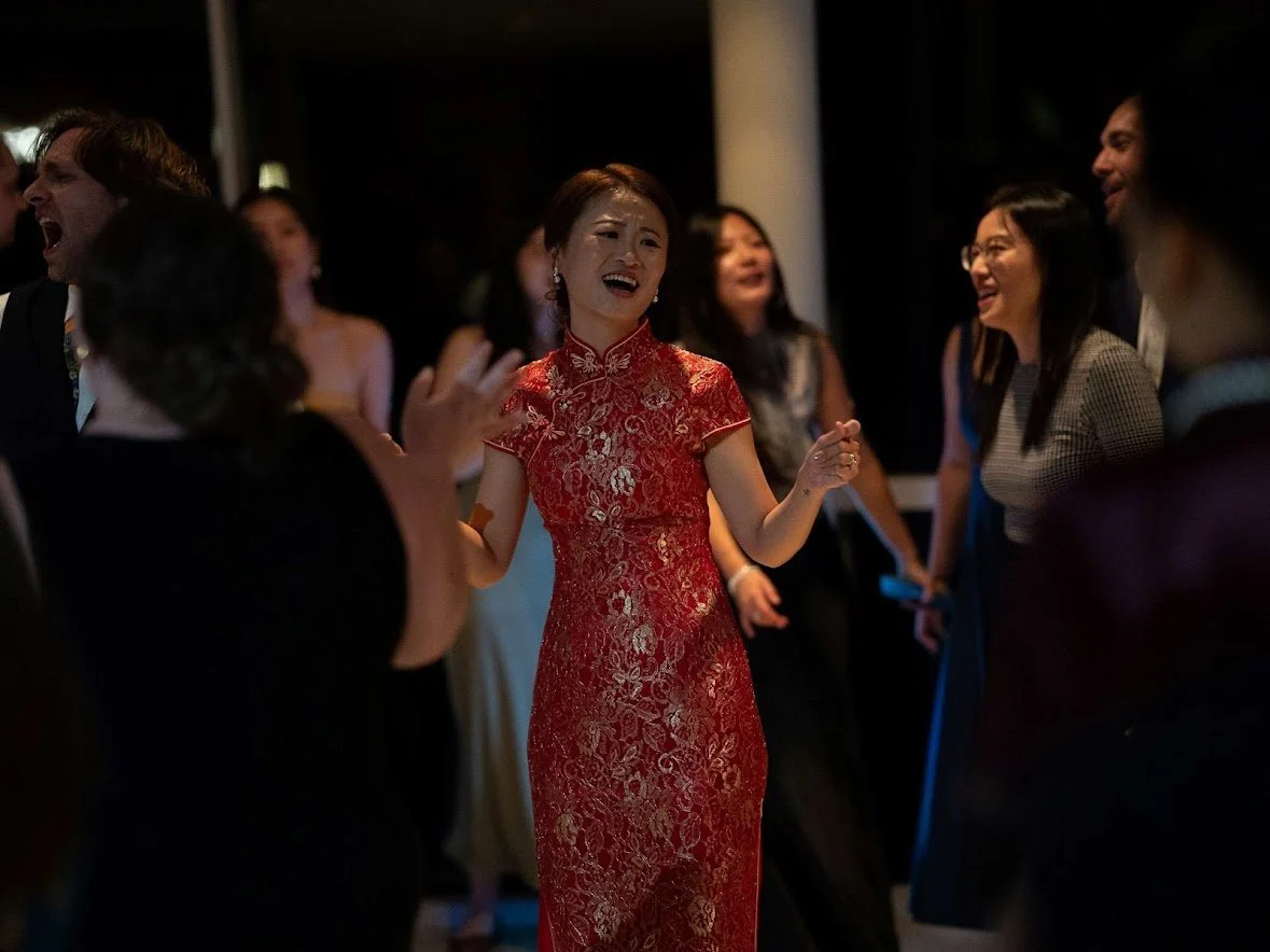 A group of people dancing and celebrating at a party, with a woman in a red lace dress smiling at the center.