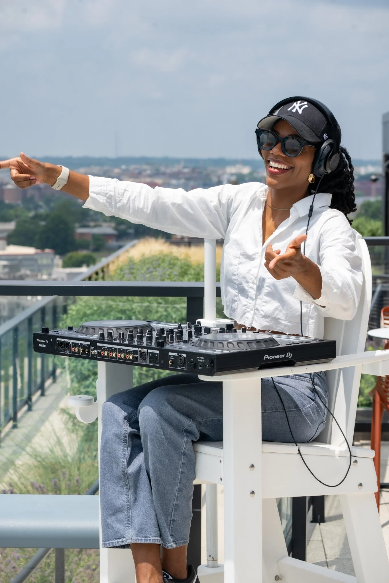 A woman smiling and wearing sunglasses, a NY baseball cap, and a white shirt, DJing outdoors on a rooftop with a city skyline in the background.