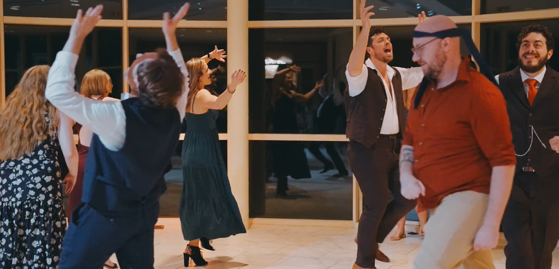 People dancing and celebrating at an indoor event, some wearing formal attire and one person dressed in a casual shirt and glasses with a headband.
