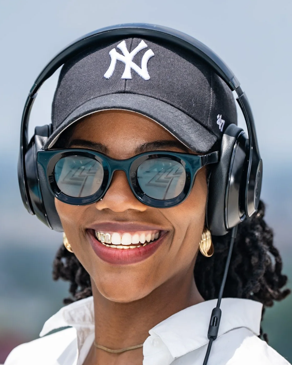 DJ Kyn, A woman wearing a black baseball cap with a white New York Yankees logo, large headphones, and sunglasses, smiling outdoors.