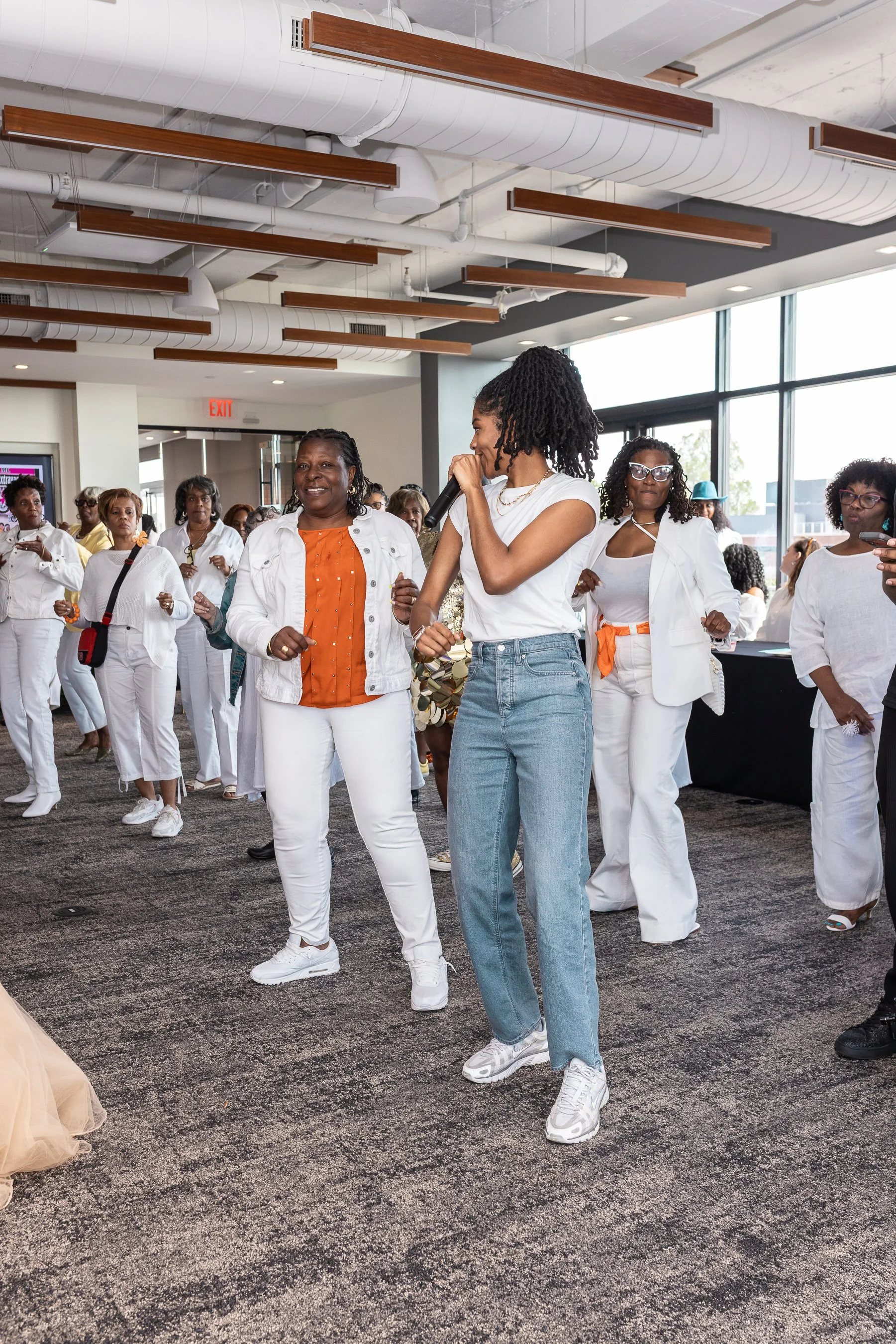A group of women dressed in white at a birthday party, with one woman holding a microphone, gathered in a modern indoor space with large windows and an industrial ceiling.