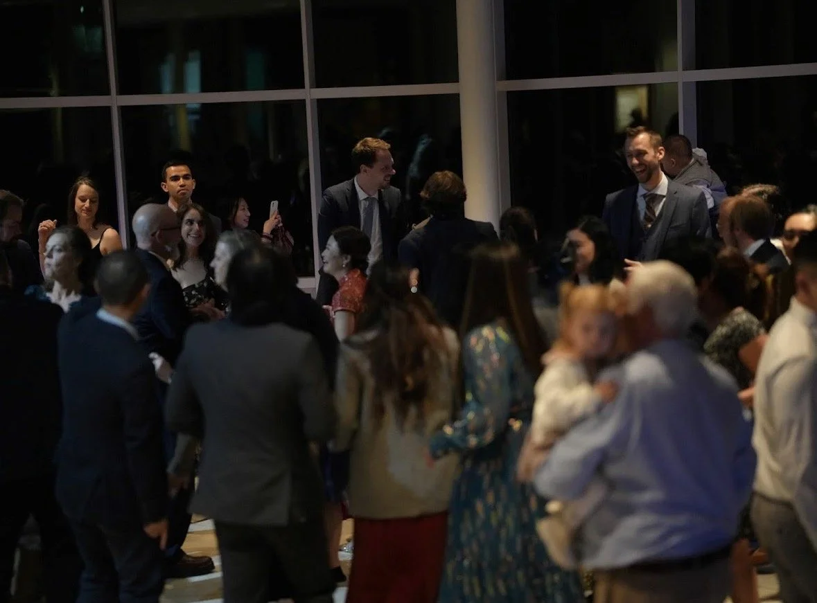 A large group of people dressed in formal attire gathered at a social event, talking and mingling indoors near large windows.