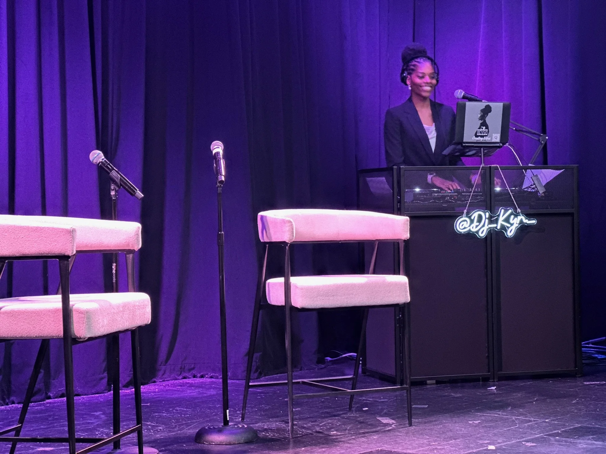 A woman with braided hair smiling while standing behind a DJ console on a stage with purple curtains, with microphone stands and pink chairs nearby.
