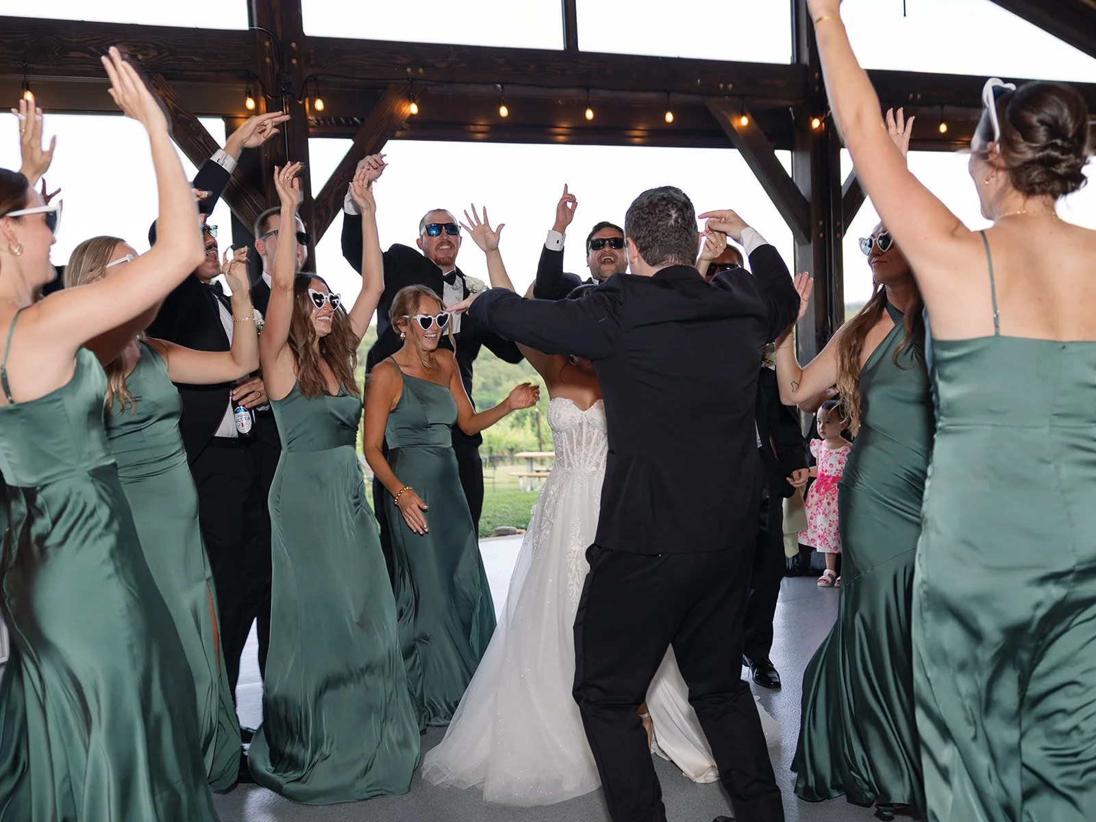 Wedding reception celebration with bride and groom dancing surrounded by bridesmaids and groomsmen, some wearing sunglasses, in a decorated venue with outdoor scenery visible through large windows.