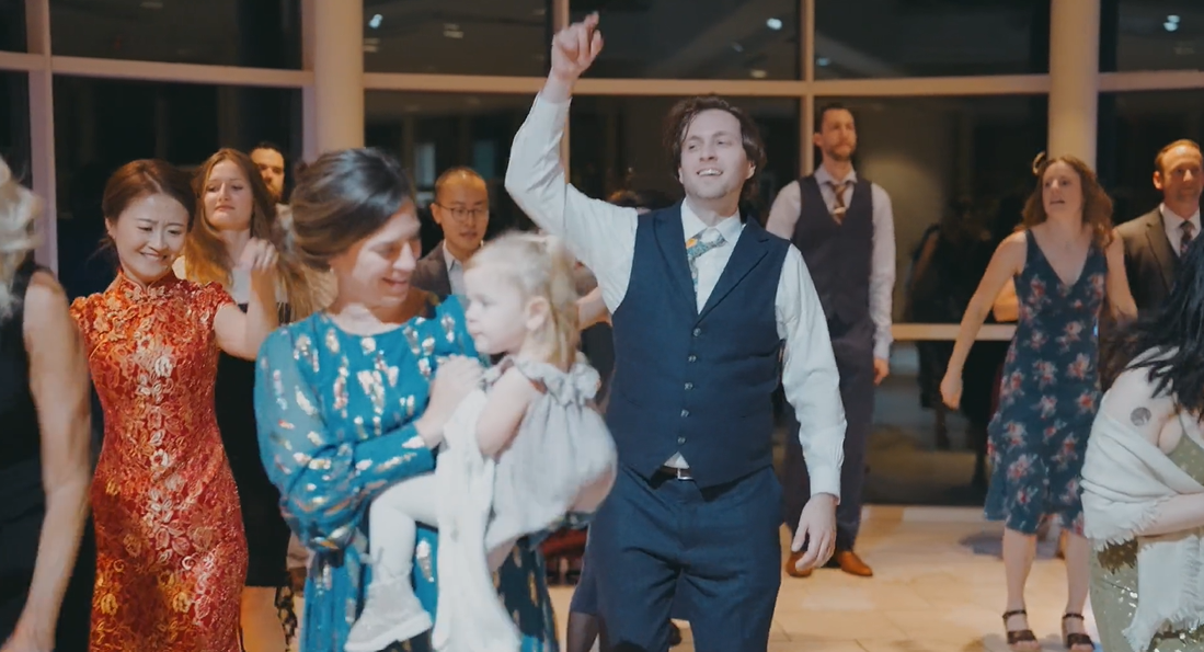 Group of people dancing at a celebration, with a woman holding a young girl and a man raising his arm in the air.