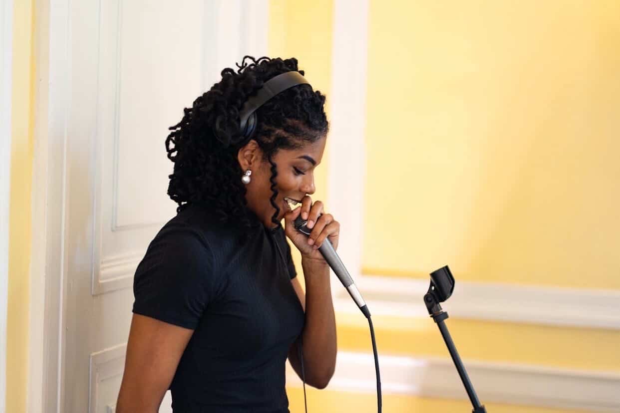 A woman with curly black hair in a black shirt wearing headphones, holding a microphone, singing or speaking into it with a smile.