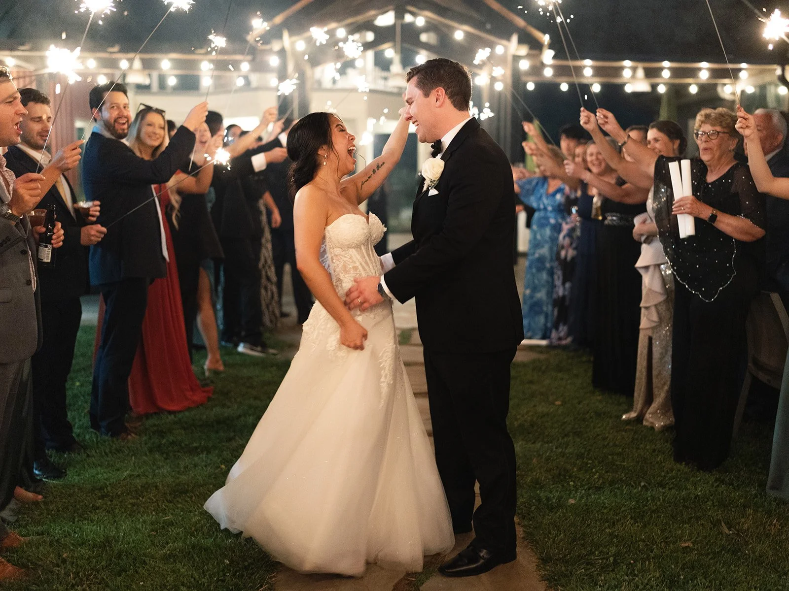 A bride and groom dancing at their wedding reception at night, surrounded by guests holding sparklers and celebrating outdoors.