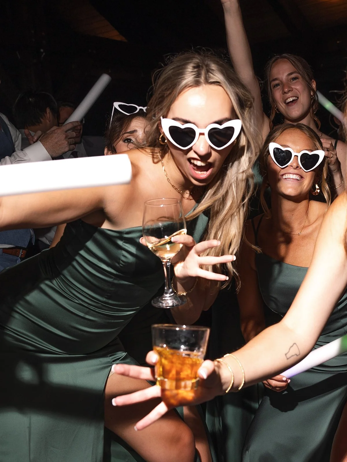 Group of women celebrating at a wedding, wearing heart-shaped sunglasses, holding wine glasses, and smiling.