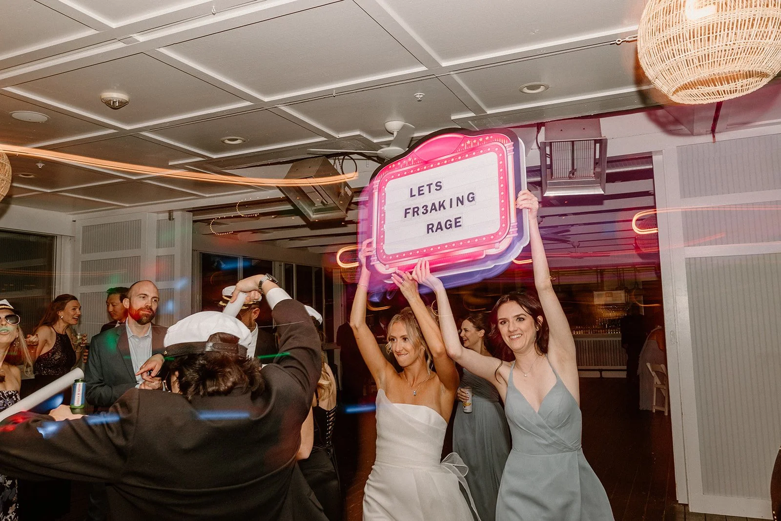 People celebrating at an indoor party with one person holding a neon sign that reads 'LETS FR3AKING RAGE' and others dancing and smiling.