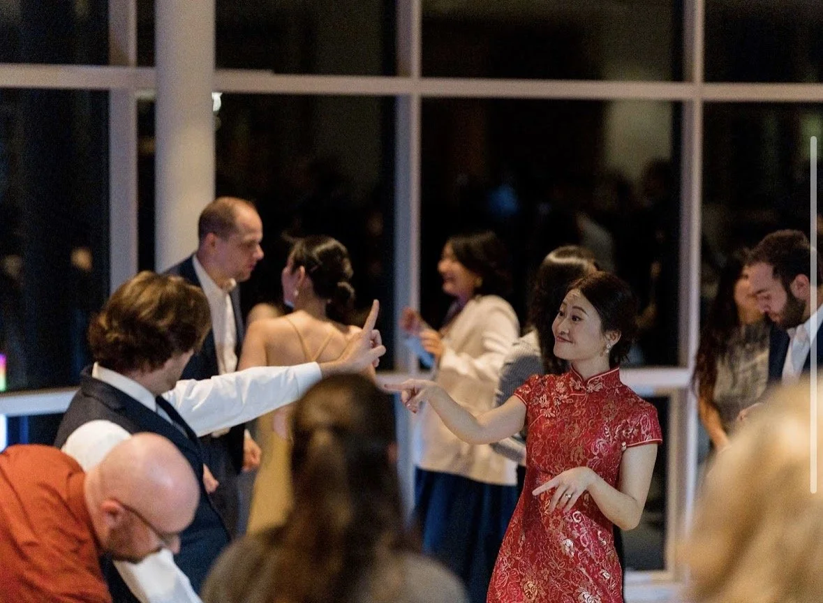 A woman in a red cheongsam dress appears to be playfully finger-pointing at a man with longer curly hair and a vest, at a wedding. People in the background are engaged in conversations.