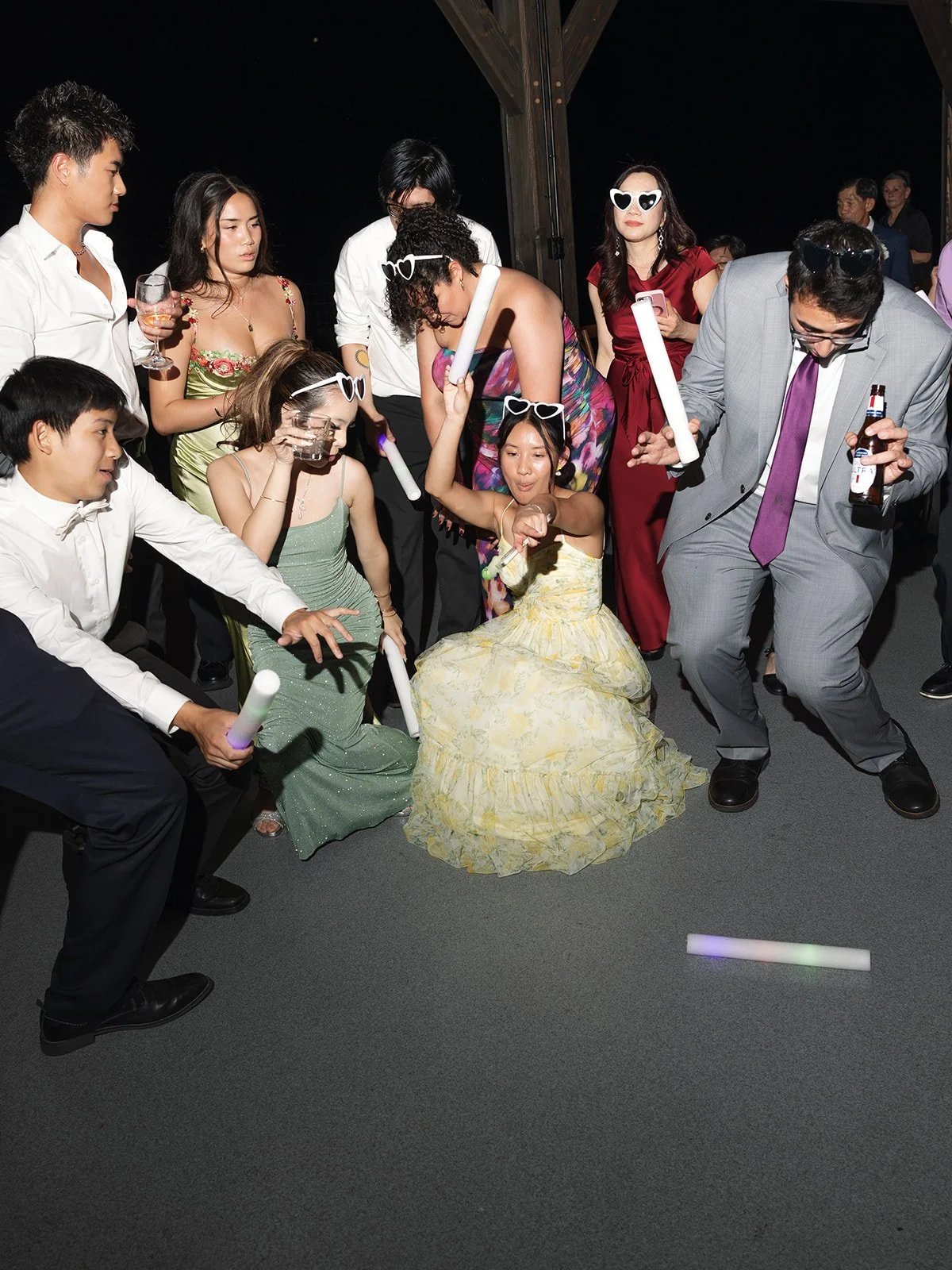 People dancing and celebrating at a party, some wearing sunglasses, holding glow sticks, and drinks, with a woman in a yellow dress in the center.