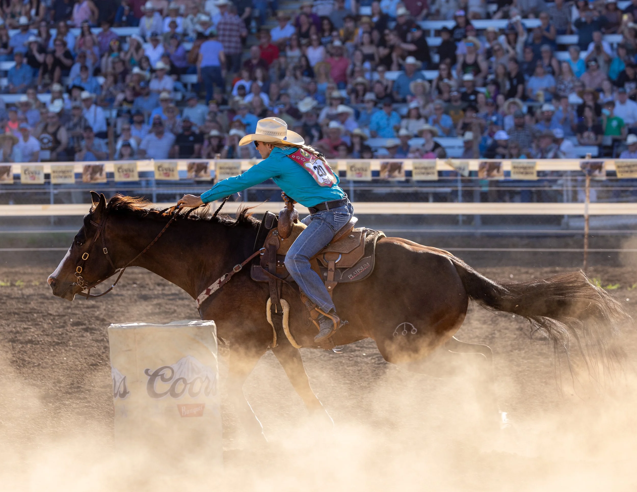 A rodeo event showing a cowboy or cowgirl riding a bucking horse on dirt ground in front of a crowd at an outdoor arena, with some spectators wearing cowboy hats.