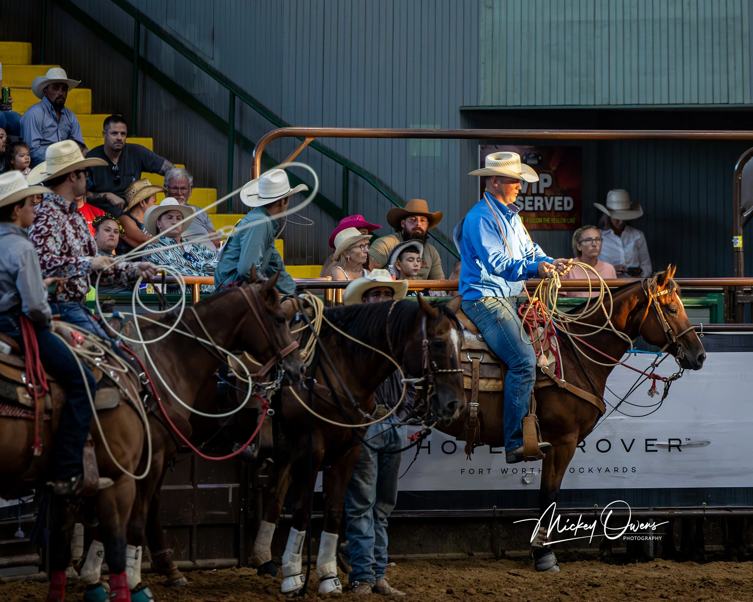 A group of people dressed in cowboy hats and western attire attending an rodeo event, with some riding horses and other spectators seated in the stands.