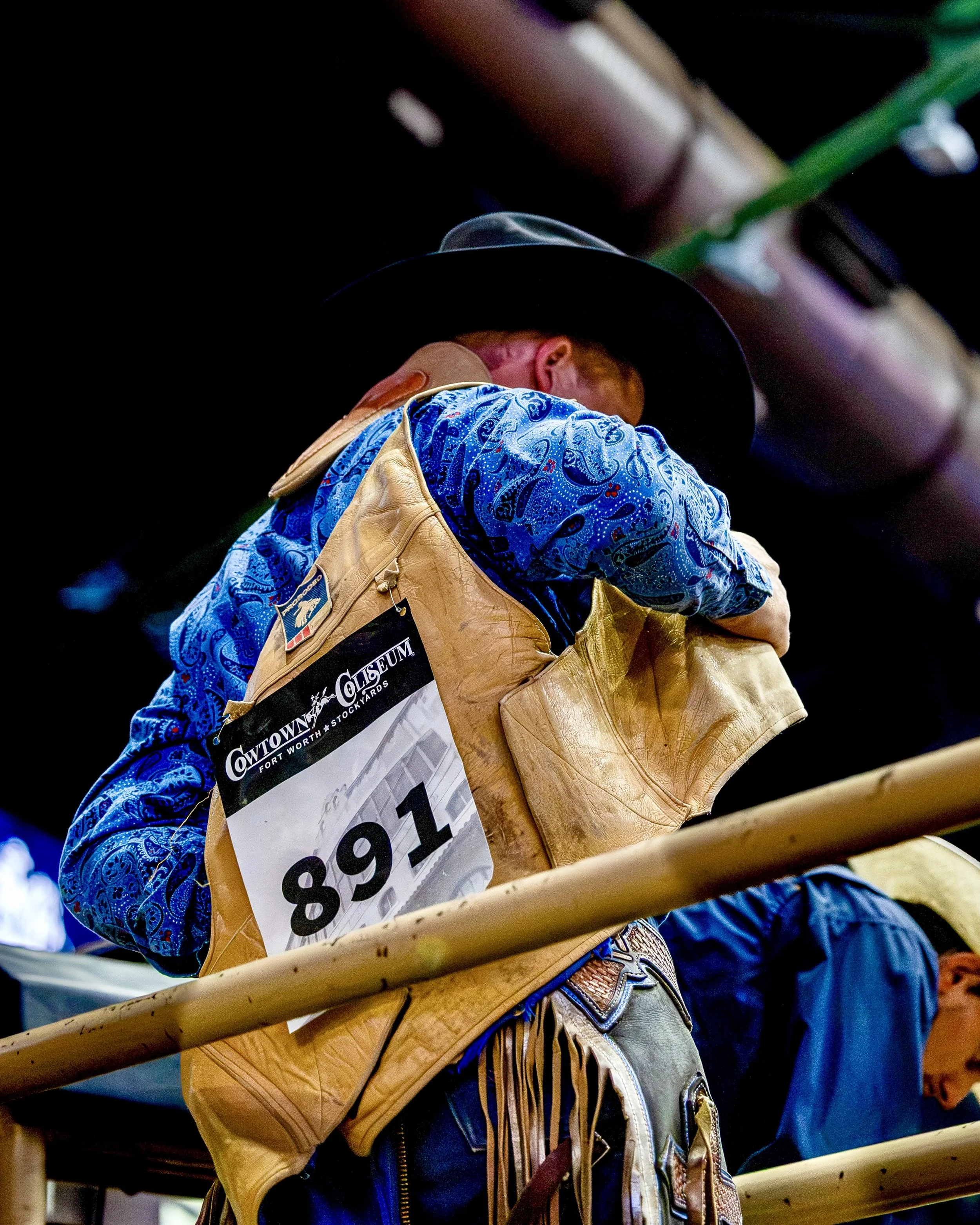 Close-up of a person wearing cowboy attire, including a fringed leather vest, patterned blue shirt, and cowboy hat, at an indoor rodeo event with a numbered badge on the vest.