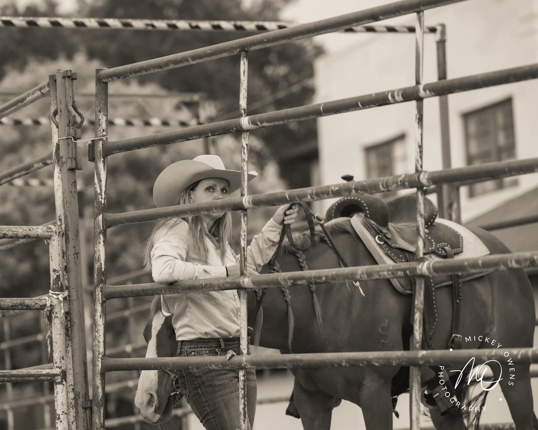 A woman wearing a cowboy hat and denim jeans leaning against a metal fence, holding a horse's bridle, with trees and a building in the background.