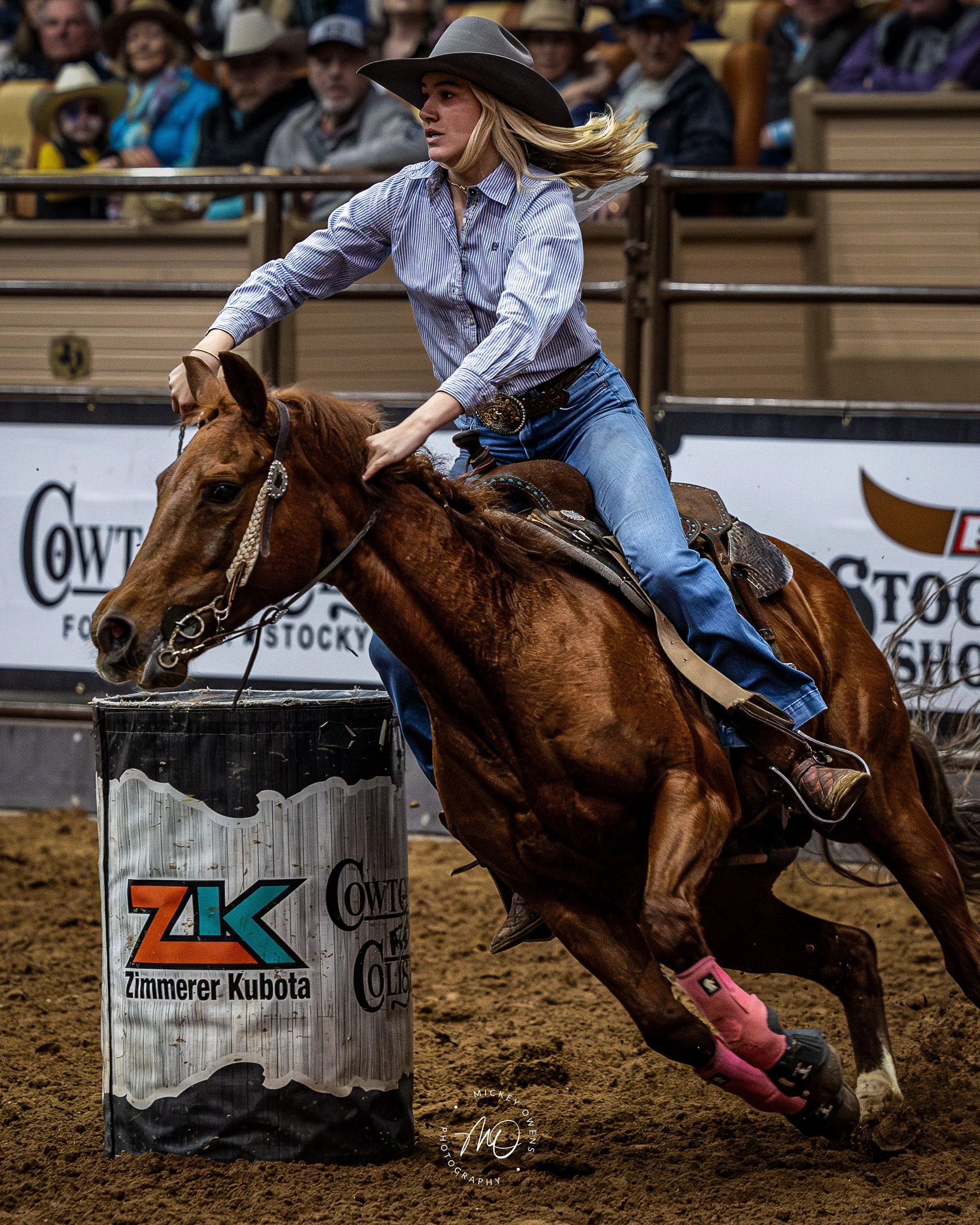 A woman riding a brown horse around a barrel during a rodeo event inside an indoor arena with spectators in the background. The woman is wearing a cowboy hat, a striped shirt, jeans, and pink protective leg gear.