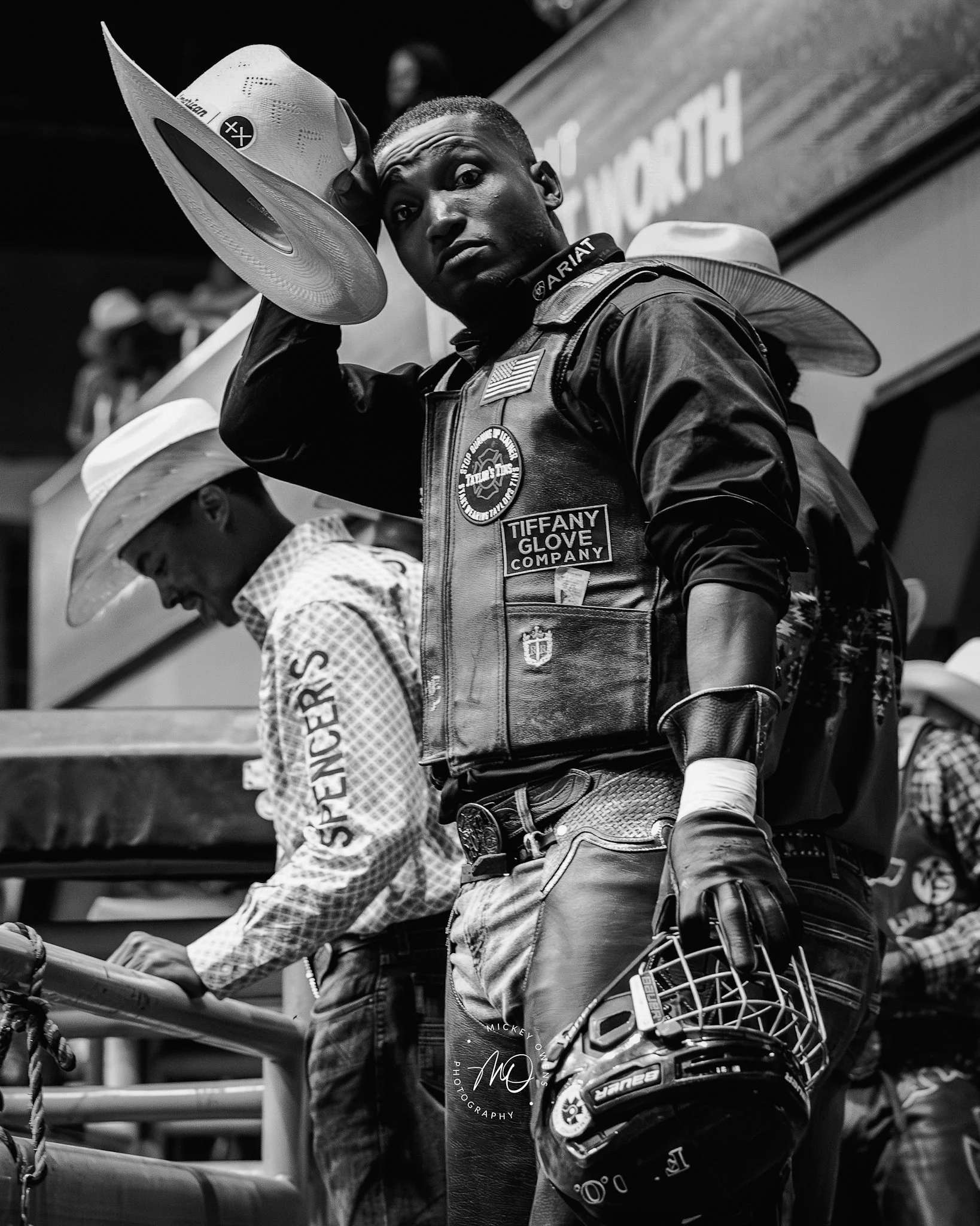 A black and white photo of a man with short hair in a racing suit, holding a hat with his right hand and looking at the camera. A woman wearing a hat and patterned shirt is in the background.