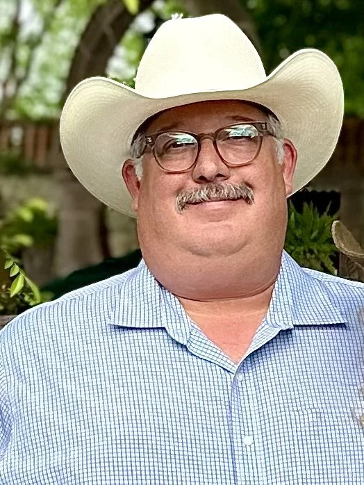 Man wearing a white cowboy hat, glasses, a light blue checkered shirt, and smiling outdoors.