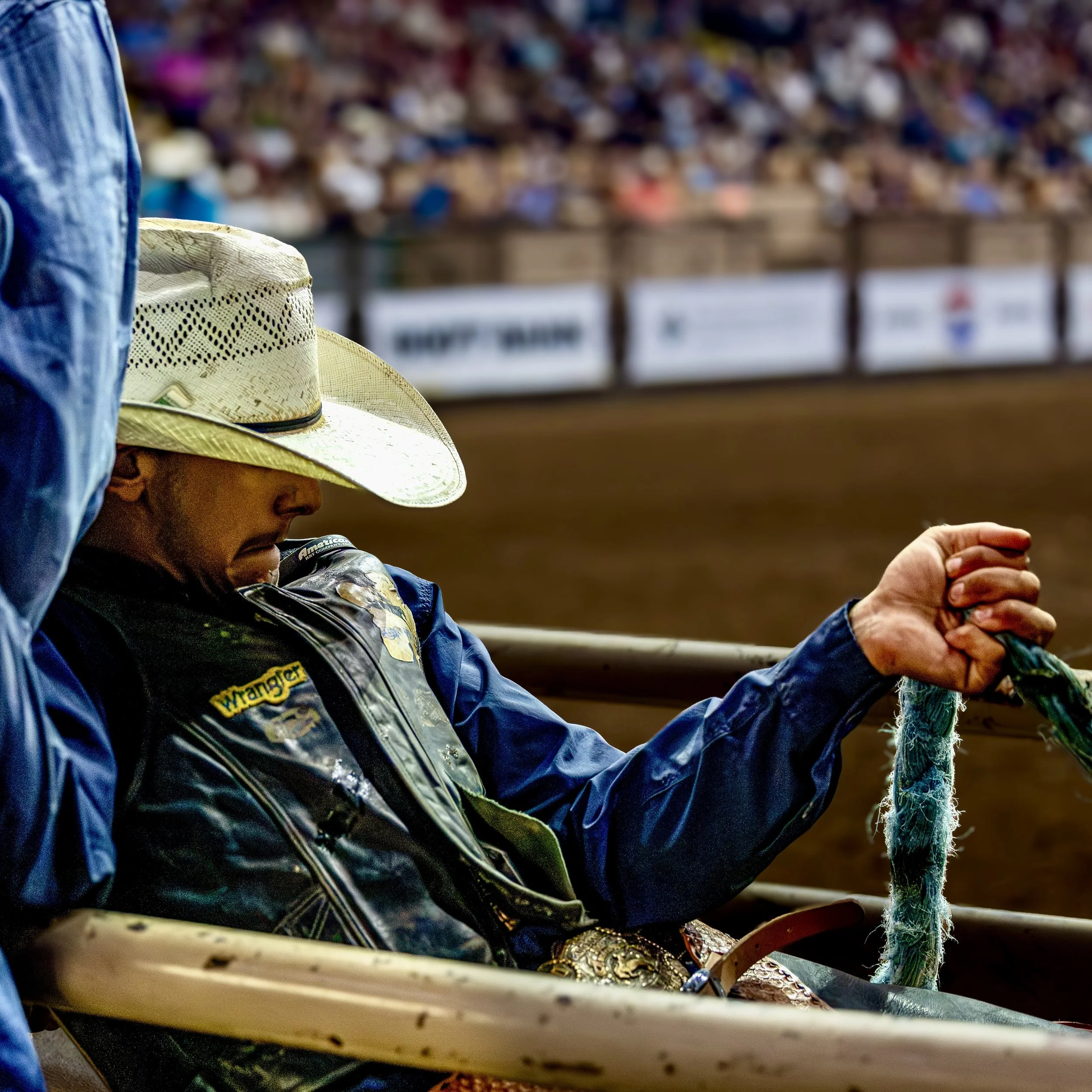 A person wearing a cowboy hat and a dark jacket with patches, sitting at a rodeo or bull riding event, with a large crowd in the background.