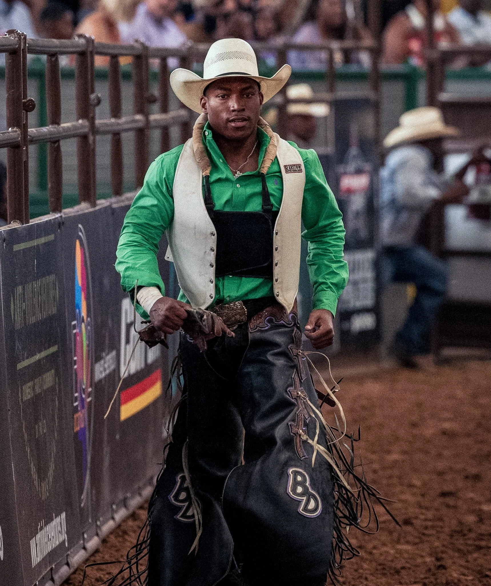 A man wearing a cowboy hat, green shirt, and black leather chaps walking in an arena with people watching in the background.