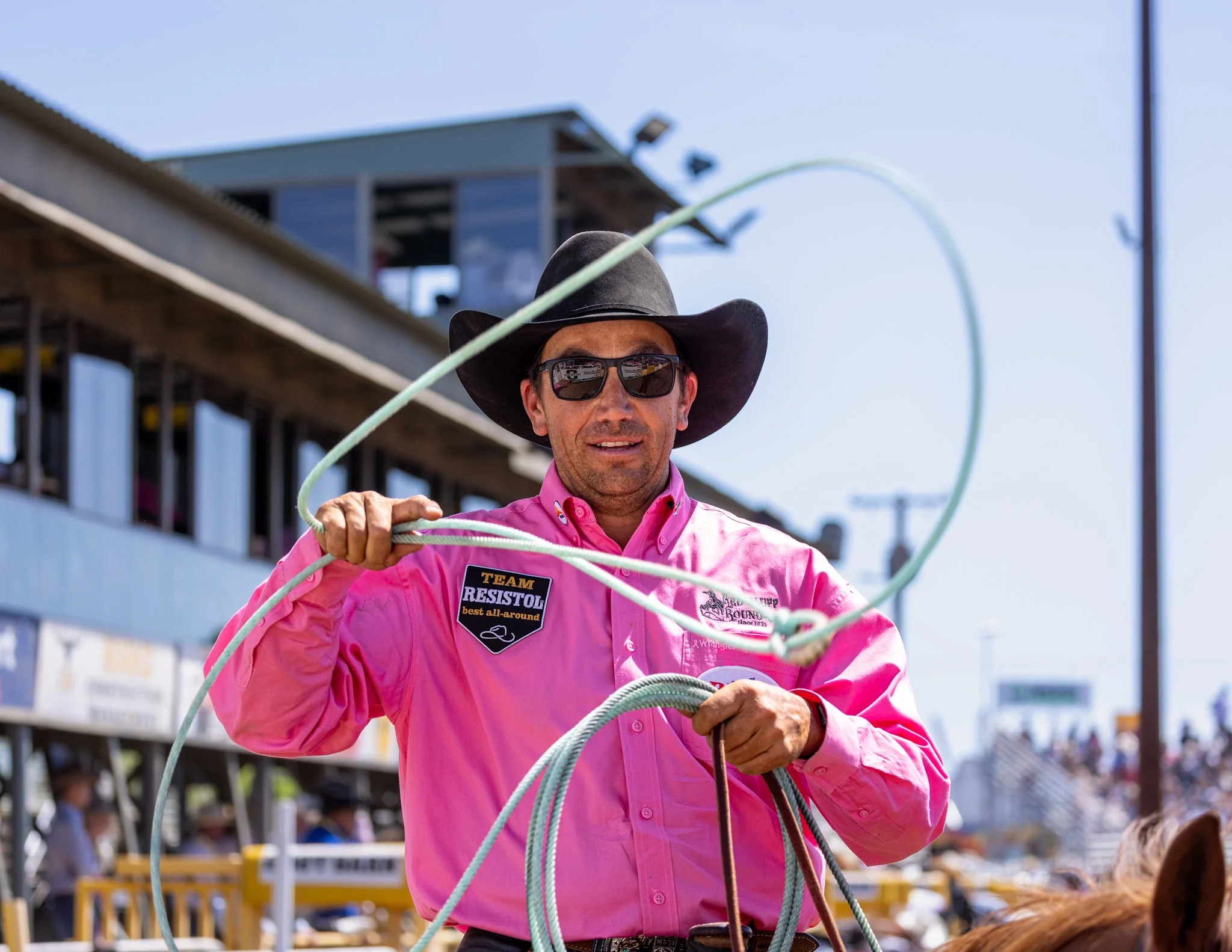 A man wearing a pink shirt, sunglasses, and a black cowboy hat, holding a rope at an outdoor event with a crowd and a building in the background.