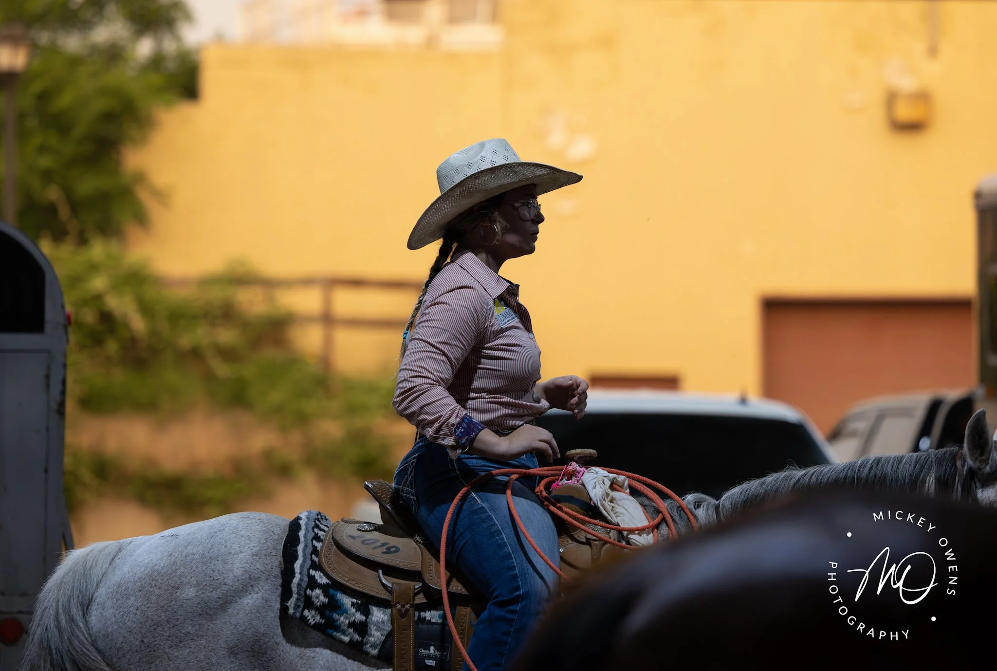 A woman riding a horse with a white coat, wearing a wide-brimmed hat, sunglasses, and a striped shirt, against a yellow building background.