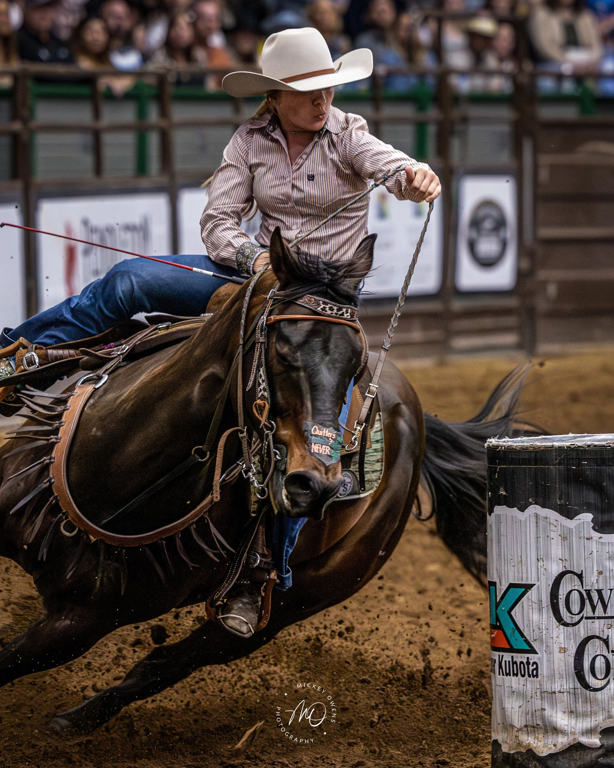 A person riding a horse in a rodeo arena, wearing a white cowboy hat, patterned shirt, and jeans, surrounded by a crowd of spectators, with a banner and a sponsor sign visible in the background.