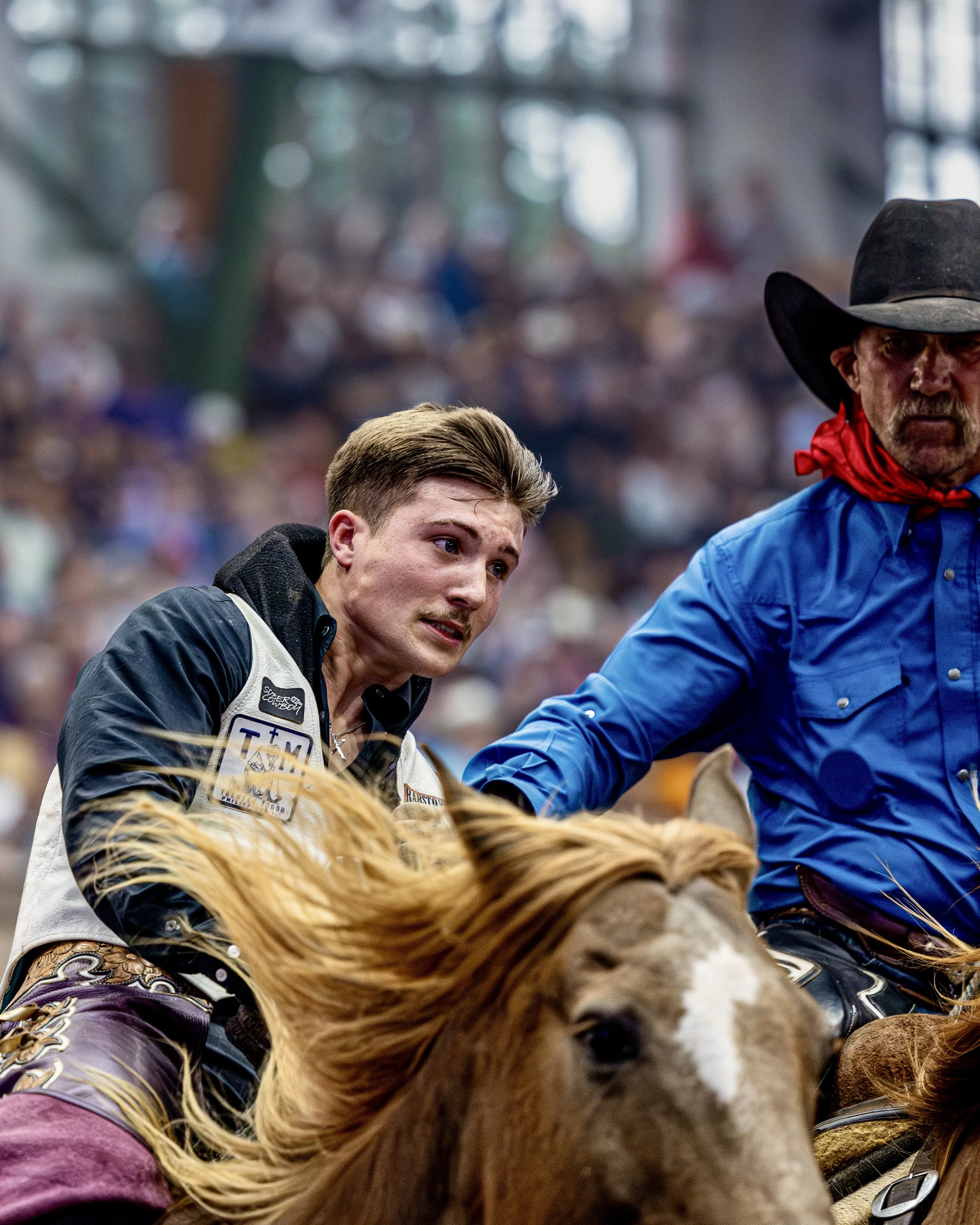 A young man and an older man with a horse in a rodeo arena, with the young man wearing a black vest, and the older man wearing a cowboy hat and red bandana. The horse has a brown mane and is wearing western tack and clothing.