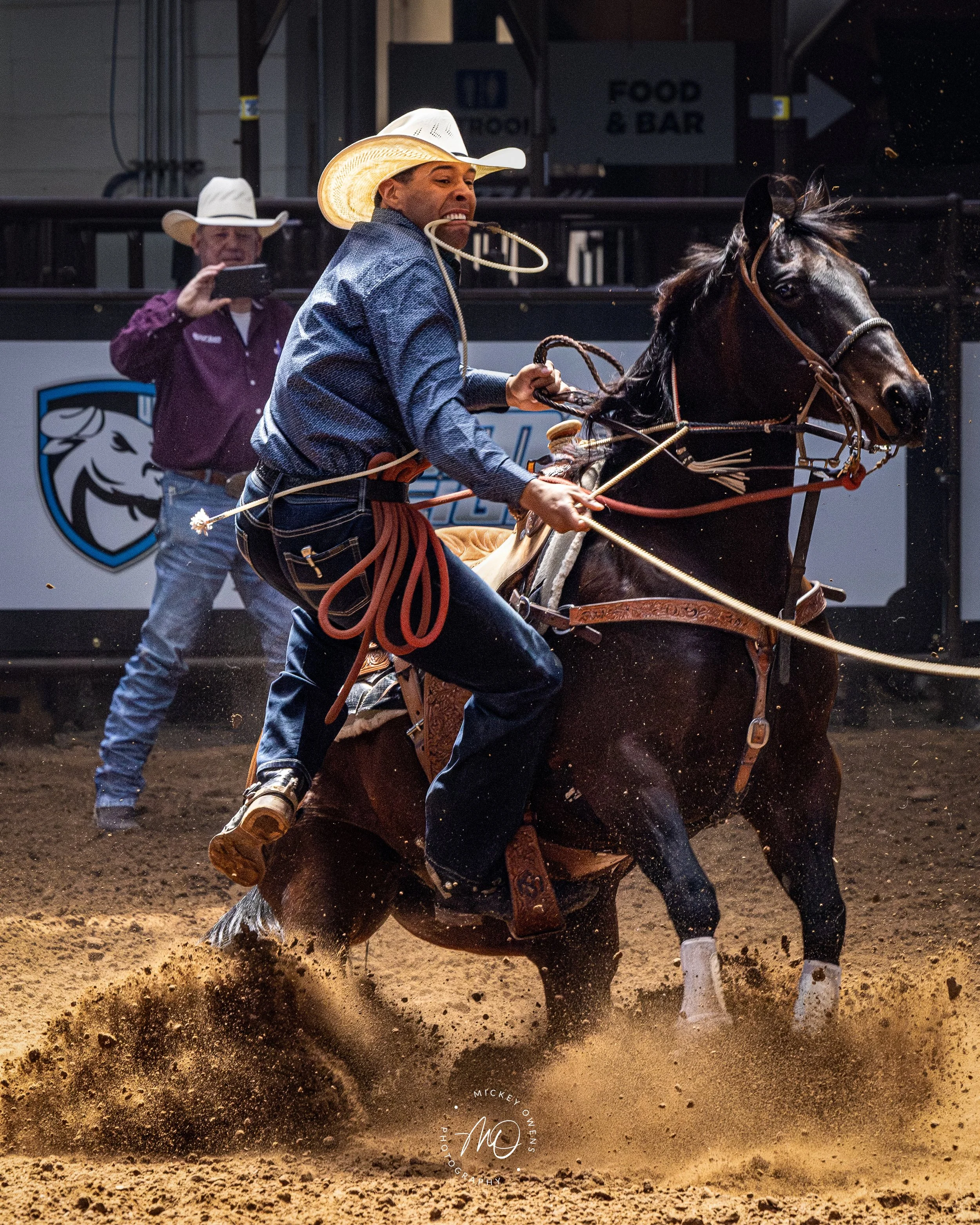 A cowboy riding a bucking bronco in an indoor rodeo arena, with another man in the background recording the event on his phone.